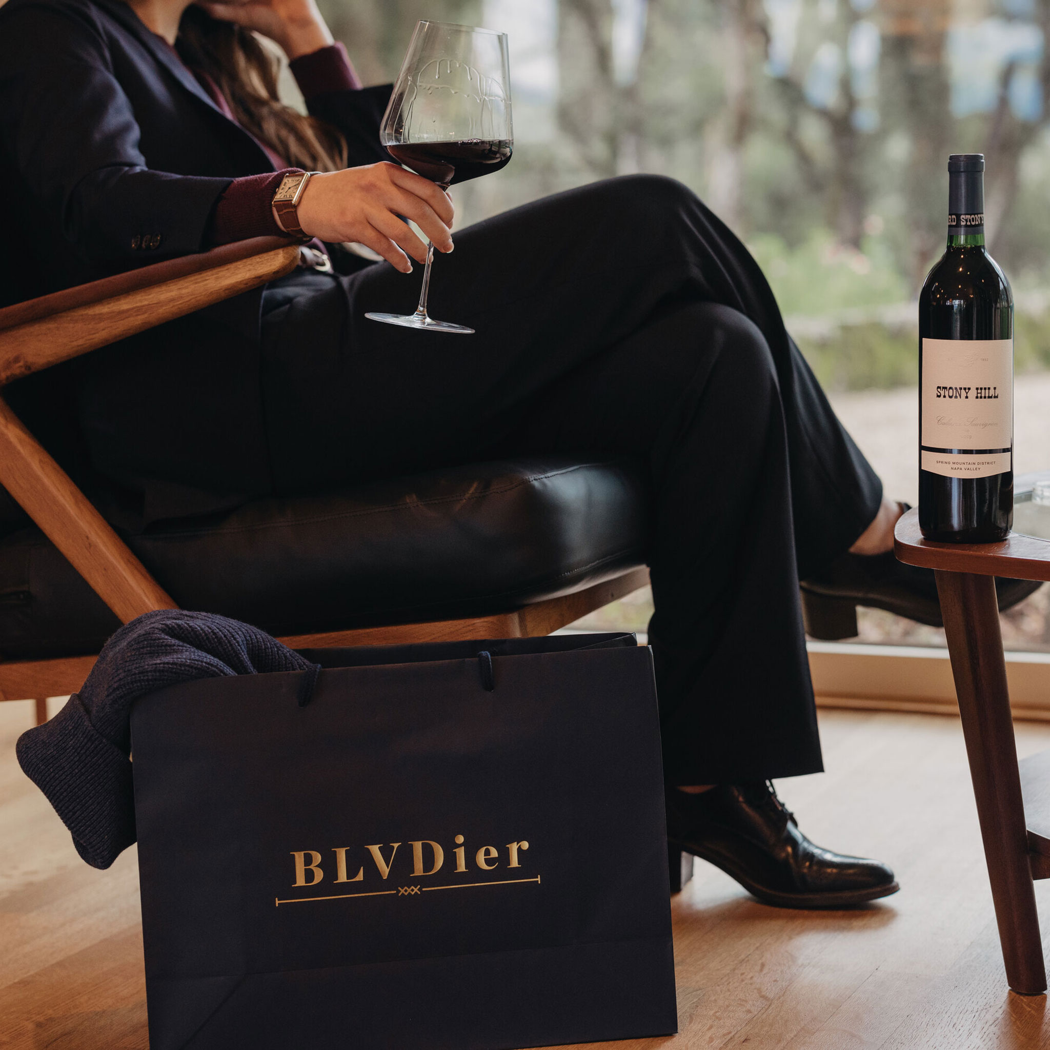 A woman holding a glass of wine while sitting in a chair next to a bag from BLVDier and a bottle of Stony Hill Cabernet Sauvignon on a table.